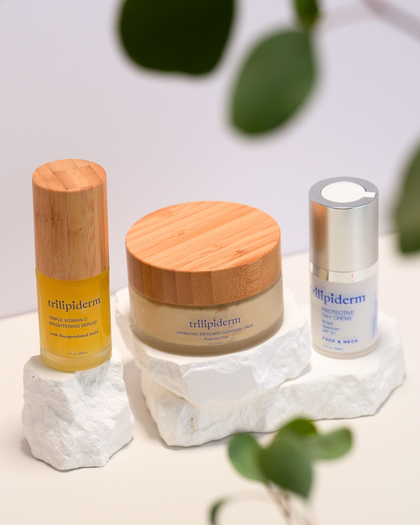 Three skincare products from the brand 'trillipiderm' on a white stone surface with green leaves in the background.