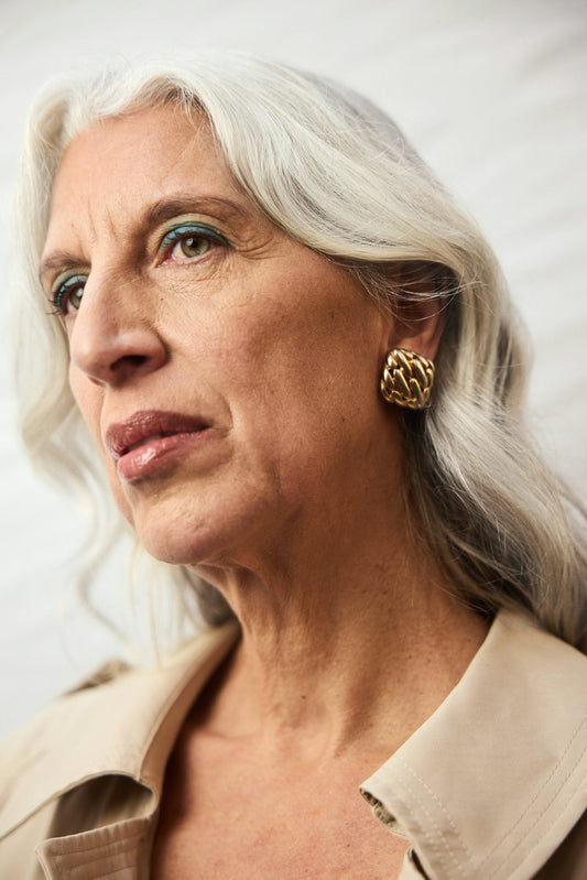 Close-up portrait of a mature woman with silver-white hair, teal eyeshadow, gold chain-link clip earrings, and visible age spots and dark spots on her skin, wearing a beige trench coat, gazing upward against a soft grey background.