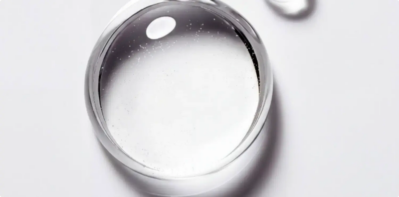 A close-up studio shot of a clear, transparent cosmetic droplet or gel sphere resting on a white surface, with soft shadows and a subtle reflection.