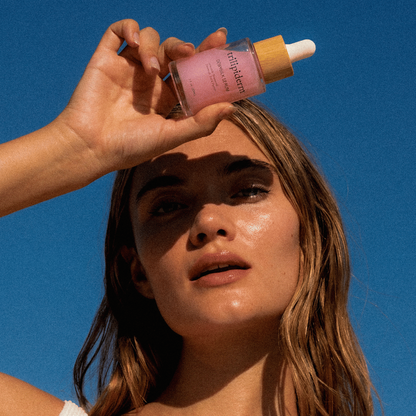 Woman holding a pink dewy silk serum with niacinamide for healthy glowing, bouncy skin. Beautiful blonde model holding dewy silk serum against and bright blue ski. Model has clear, bright even skin, that's deeply hydrated.