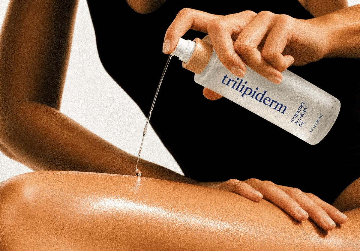 Trilipiderm Hydrating All-Body Oil poured from its frosted bottle onto a glowing, hydrated women's leg.