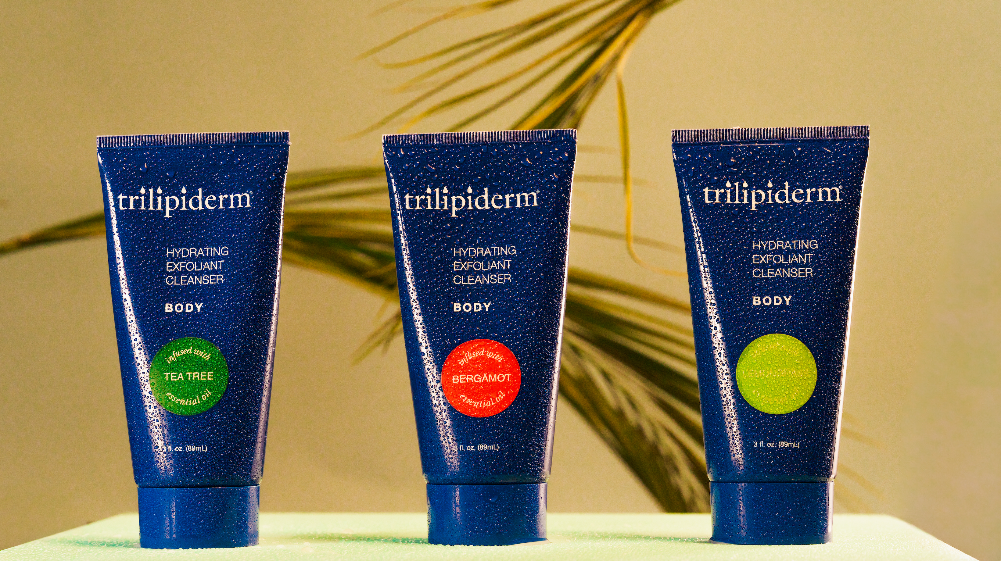 Three blue tubes of trillipiderm body cleansers with infused essential oils. on a light green surface with a blurred plant background.