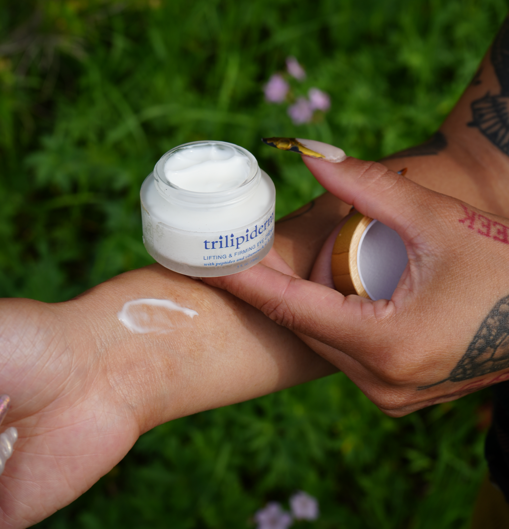 Trilipiderm Lifting & Firming Eye Crème jar held in hand with creamy swatch applied on skin, showing smooth lightweight texture against a natural green background. Hydrating eye treatment with peptides and botanical lipids to reduce puffiness, brighten under-eyes, and firm delicate skin.