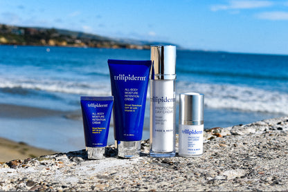 A beach setting with Trilipiderm sun protection products including a tube, bottle, and box of sunscreen, and a bottle of moisture retention cream on rock