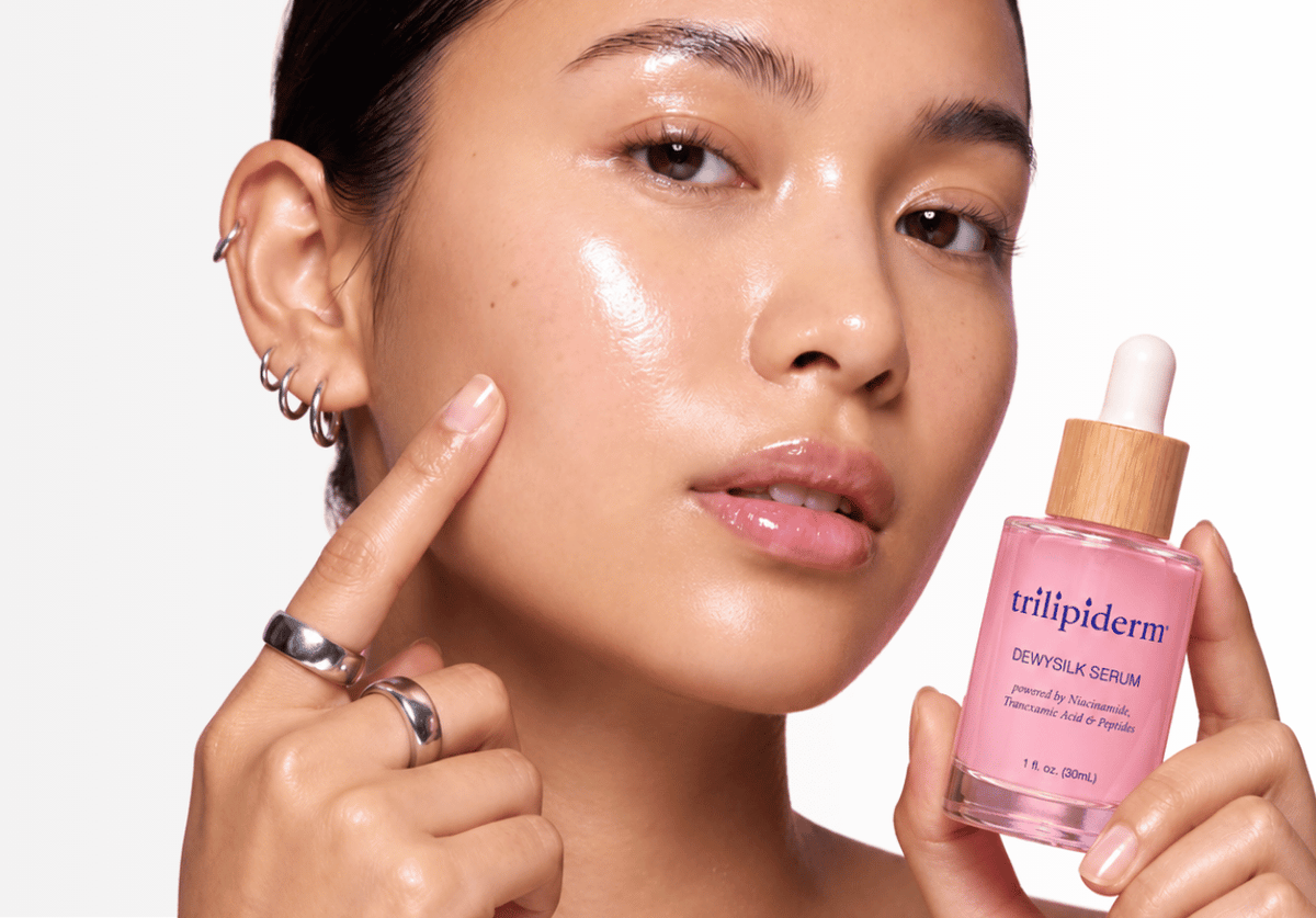 A close-up of a woman with plump, glossy, hydrated skin holding a bottle of Trilipiderm Dewy Silk peptide Serum next to her cheek, pointing to her face. The bottle is pink with a light wood dropper cap.