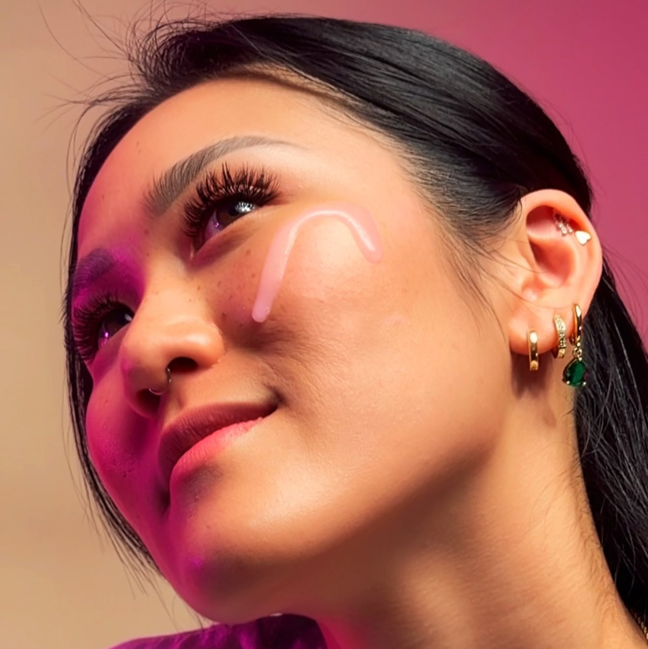 Close-up portrait of a model wearing Trilipiderm DewySilk Serum applied to the cheek, showing a glossy glass-skin glow under pink and purple studio lighting. Lightweight hydrating face serum visibly layered on smooth, radiant skin to enhance brightness, improve texture, and support barrier repair. Dewy finish highlights niacinamide and tranexamic acid brightening serum benefits for luminous, healthy-looking skin.