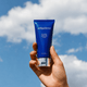 A hand holding a blue tube of trilipiderm Full Body Moisture Retention Crème against a bright blue sky with white clouds. Trilipiderm All-Body Moisture Retention Crème customer review highlighting rich hydration, improved moisture retention and softer, smoother-looking dry skin.