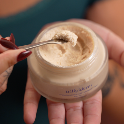 wwome holding a jar of Trilipiderm hydrating exfoliating cleanser scrub with a spoonful of scrub showing the texture of the skincare product.
