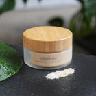Jar of hydrating exfoliating cleanser scrub  with a wooden lid on a textured surface with green leaves in the background. Showing off the texture of the scrub 