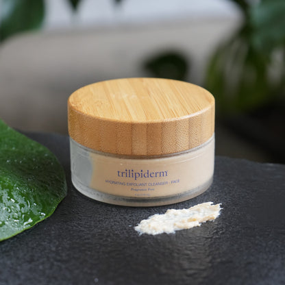 Jar of hydrating exfoliating cleanser scrub  with a wooden lid on a textured surface with green leaves in the background. Showing off the texture of the scrub 