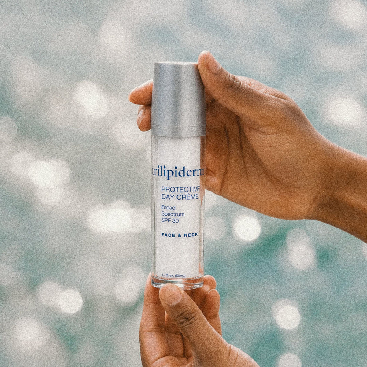 Close-up lifestyle skincare photograph of hands holding Trilipiderm Protective Day Crème Broad Spectrum SPF 30 face and neck sunscreen moisturizer, 1.7 fl. oz. (50 mL), against a softly blurred turquoise swimming pool with shimmering sunlight reflections. The sleek white bottle features a metallic silver cap and signature blue Trilipiderm branding. This lightweight daily SPF 30 moisturizer combines broad-spectrum sun protection with hydrating skincare benefits to help support the skin's skin barrier.