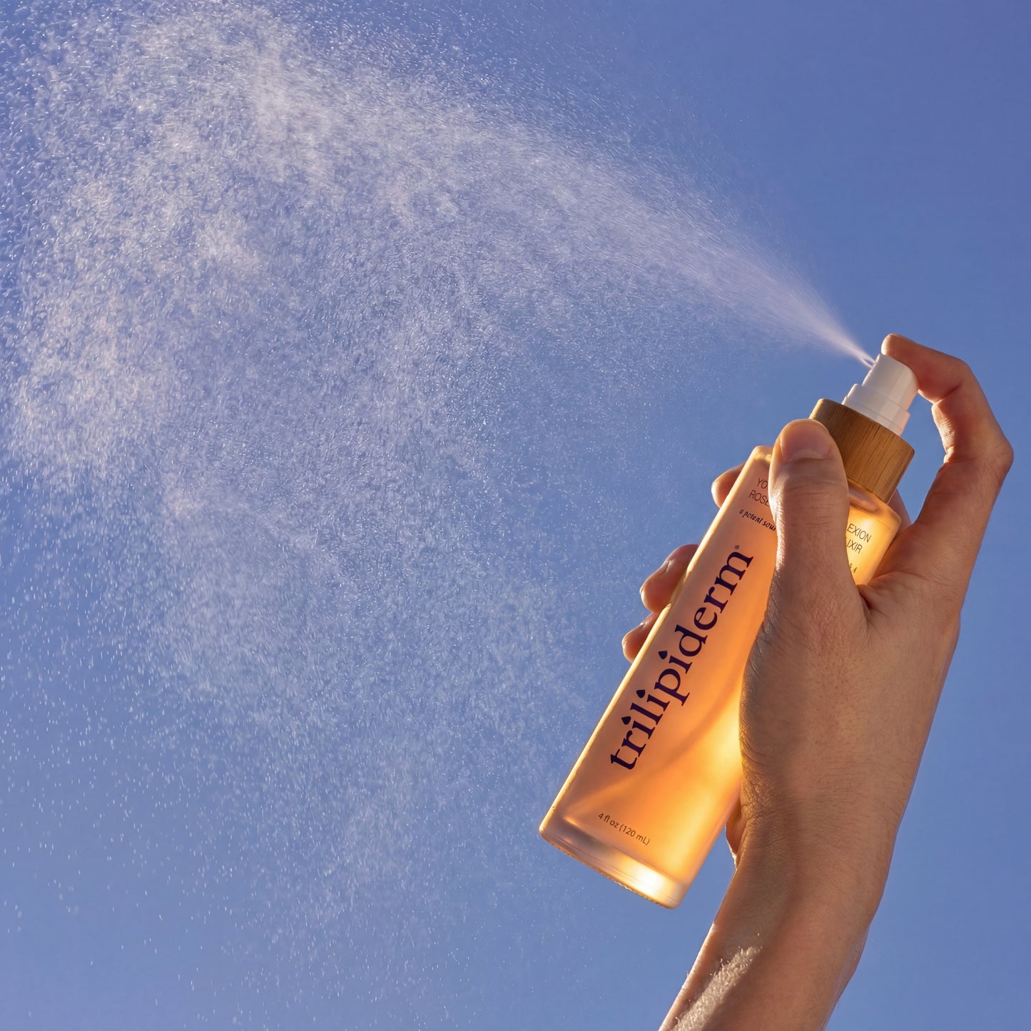 Trilipiderm Youthful Complexion Rosehip Toning Elixir being sprayed into air, capturing fine facial mist particles against blue sky lighting. Frosted blush glass bottle with bamboo collar and white spray nozzle delivering antioxidant-rich rosehip facial toner for instant hydration, barrier support, and luminous glass-skin glow. Lightweight refreshing mist suitable for all skin types.