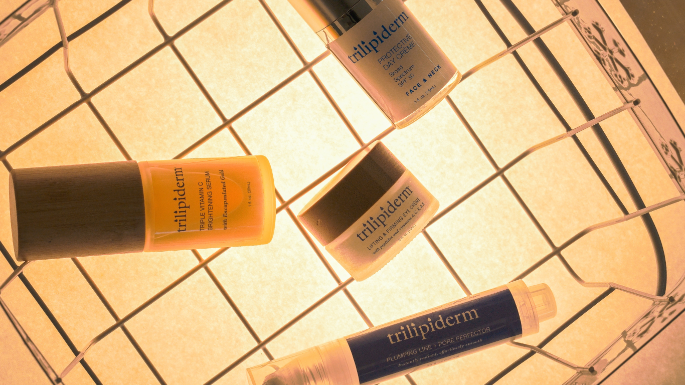 Trilipiderm USA-made serums and eye cream arranged on warm backlit surface—gold Vitamin C serum, SPF 30 day crème, firming eye crème, and plumping pore perfector for holiday skincare shoppers